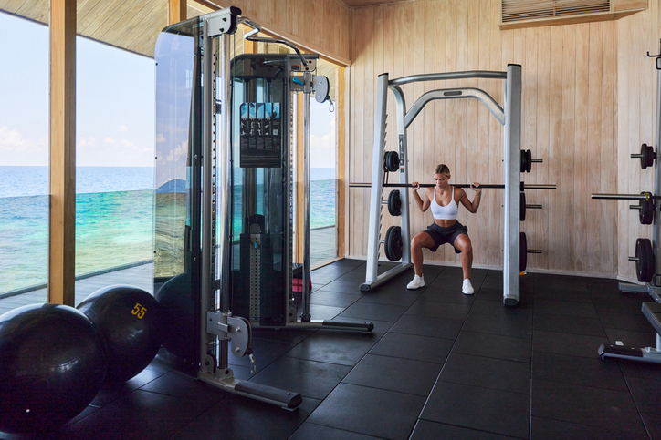 Vrouw doet squats met een halterstang in een moderne fitnessruimte met uitzicht op zee